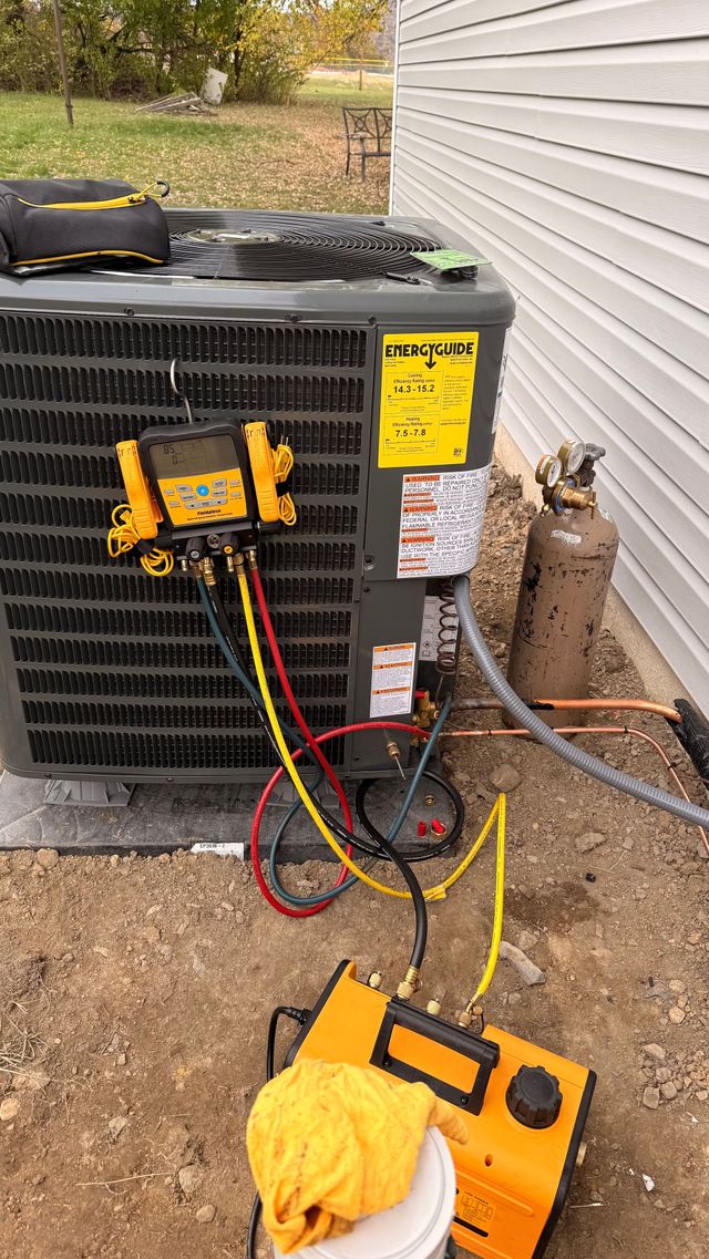 HVAC technician servicing an air conditioning unit; hoses, gauges, and a refrigerant tank are connected.