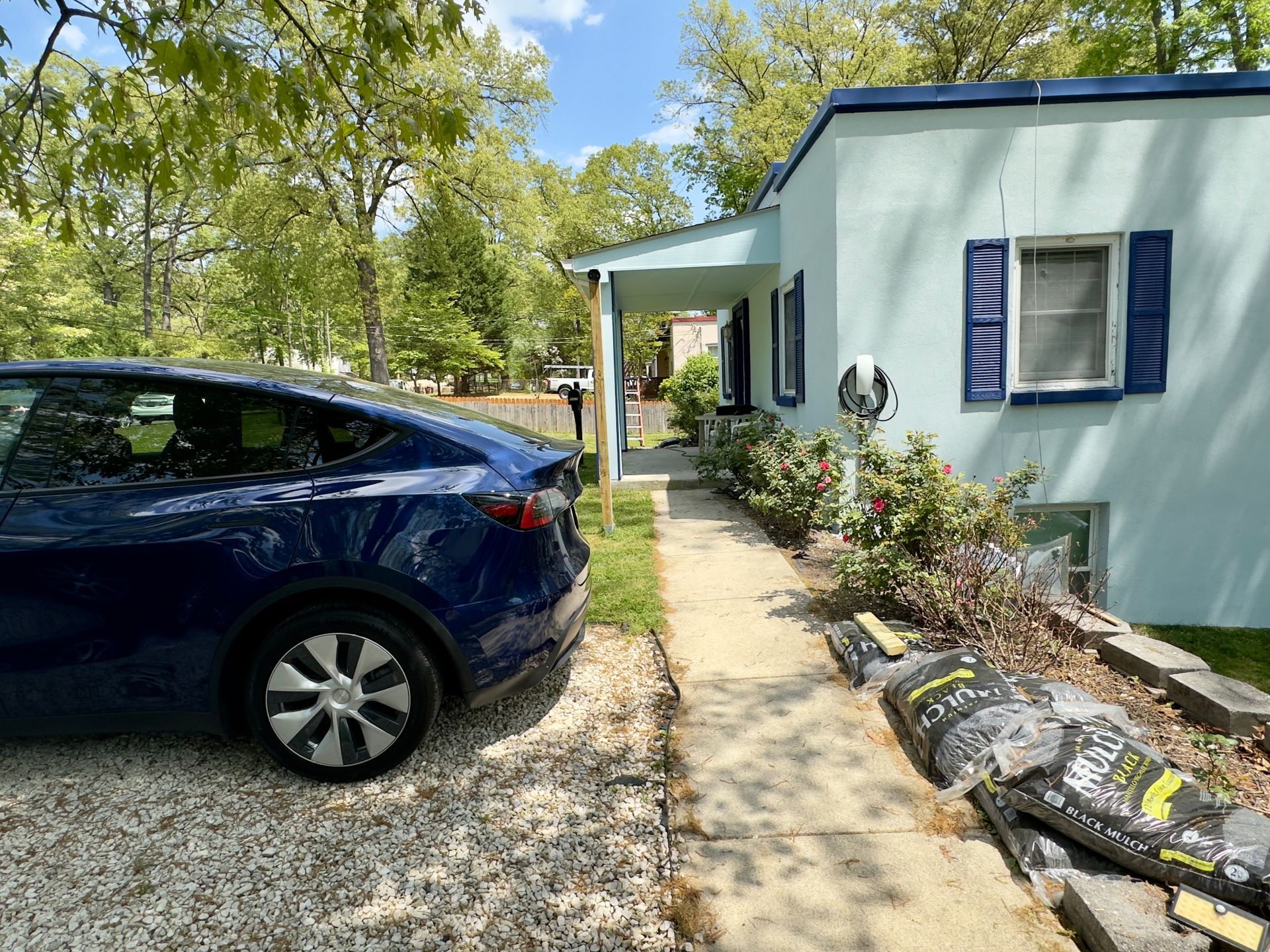 Blue Tesla parked beside a pale blue house with an EV charger; pathway leads to the entrance.