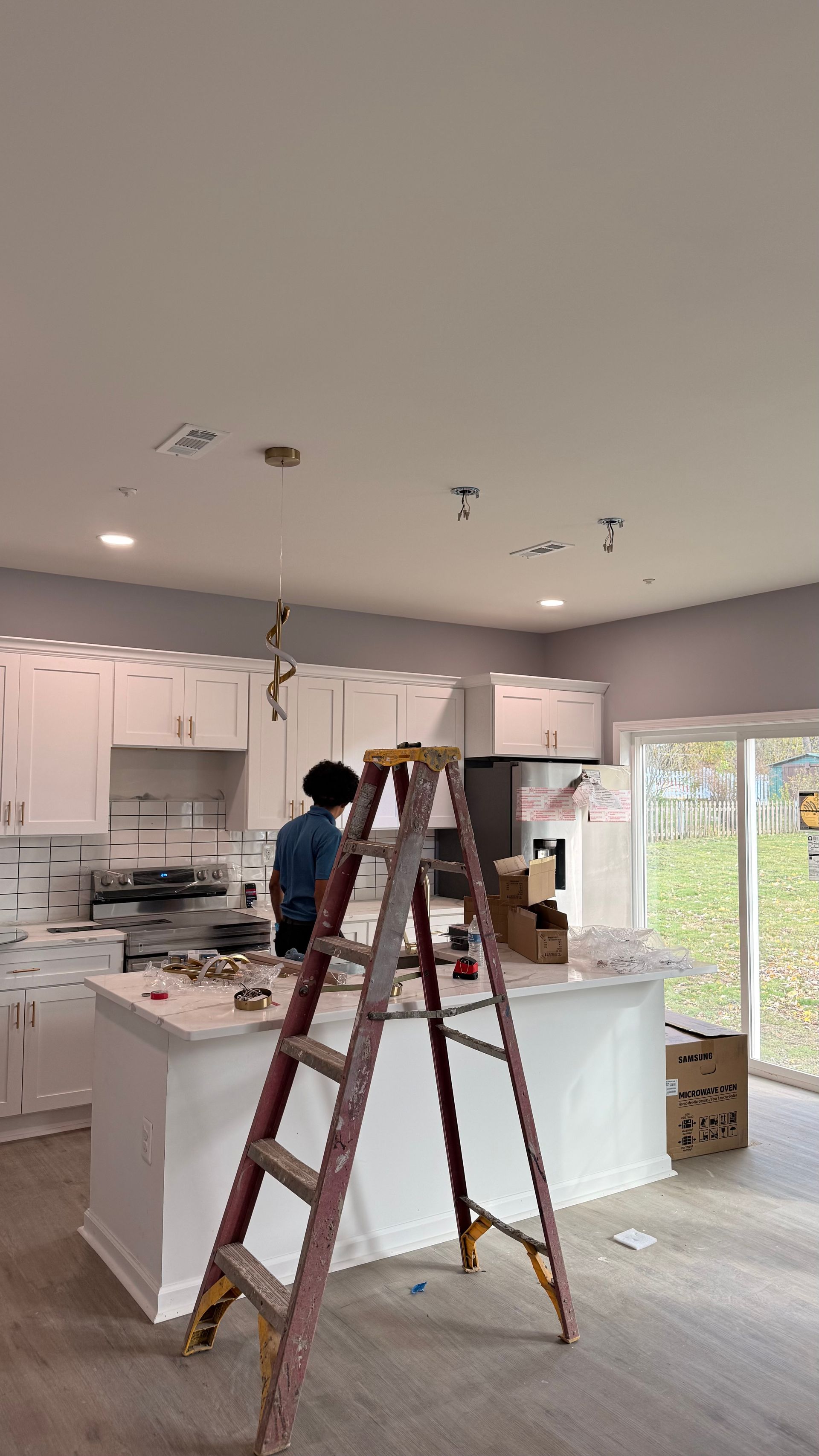Person on a ladder installing a light fixture in a modern white kitchen. Cabinets and island are visible.