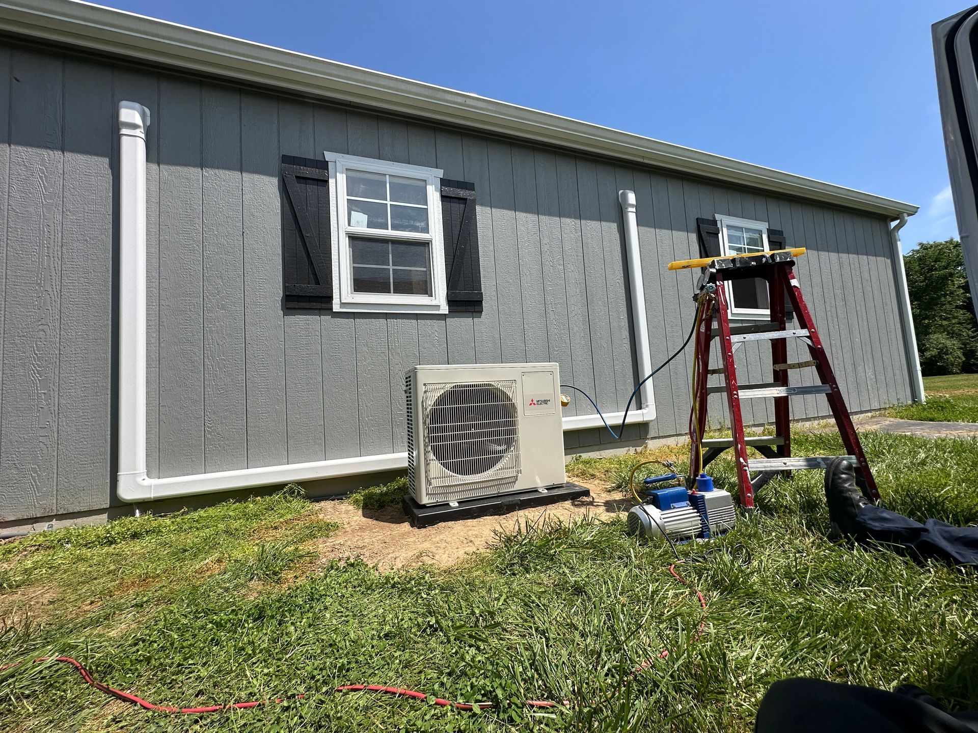 An air conditioning unit is outside a gray building with two windows and black shutters. 
