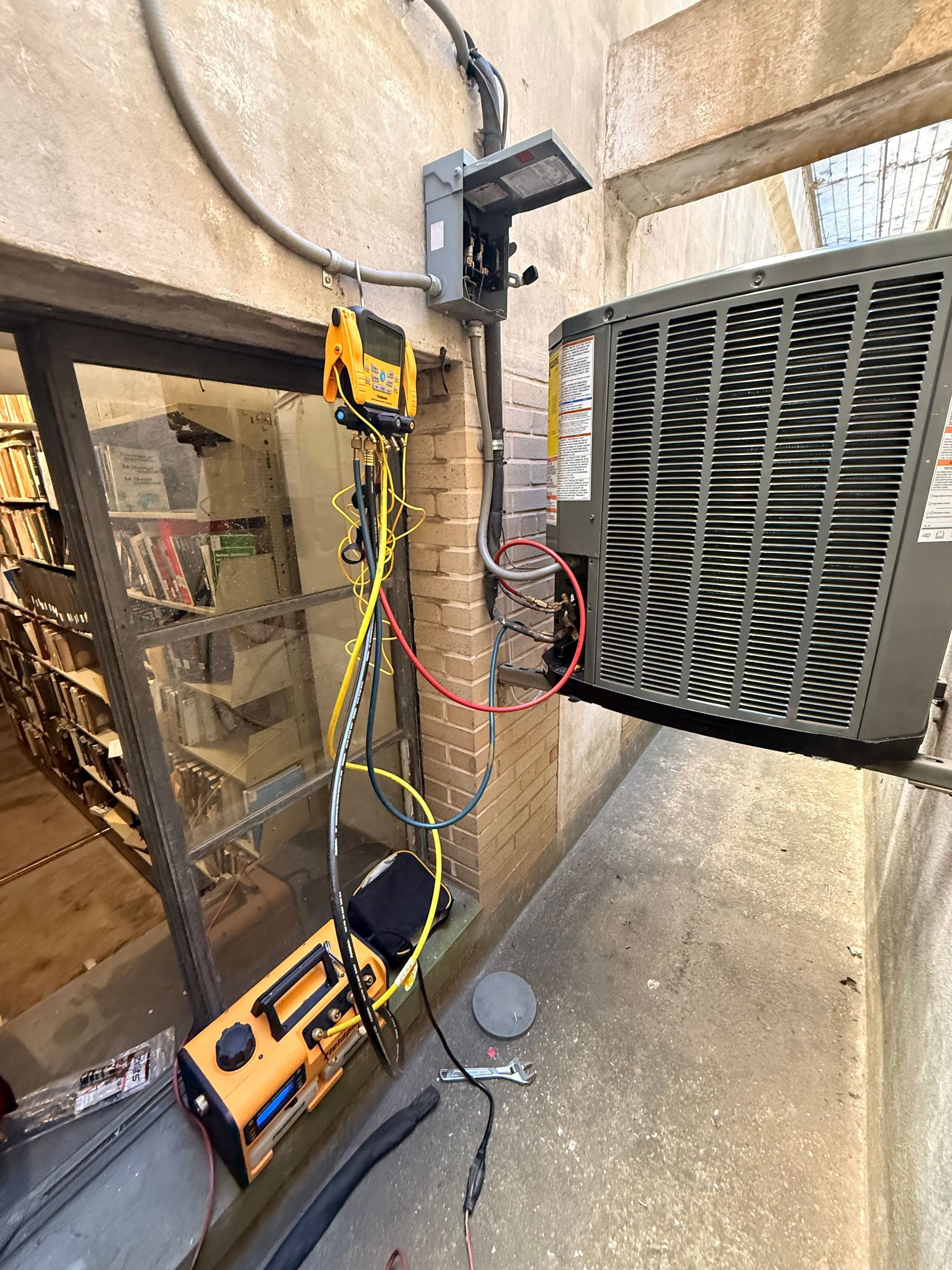 HVAC unit on brick wall with testing equipment. Yellow cables, gray and orange equipment. Library books visible.
