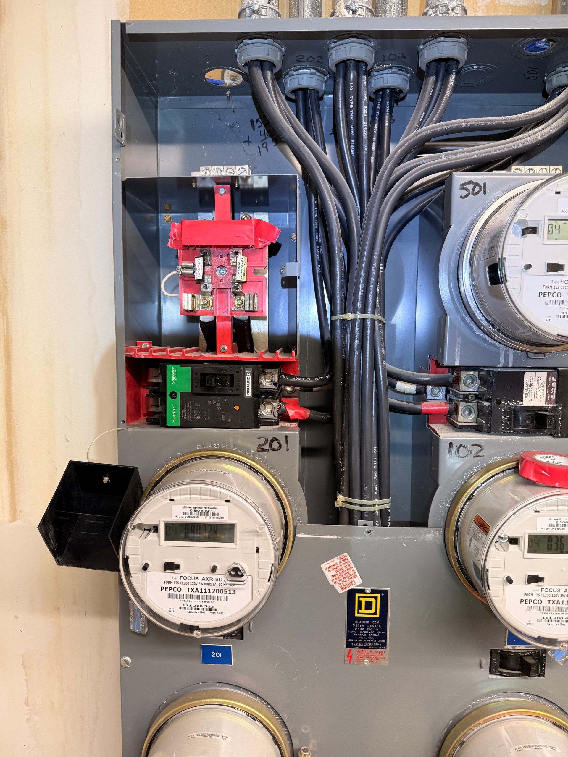 Electrical panel with multiple meters, conduits, and circuit breakers. Gray, red, and black components dominate.
