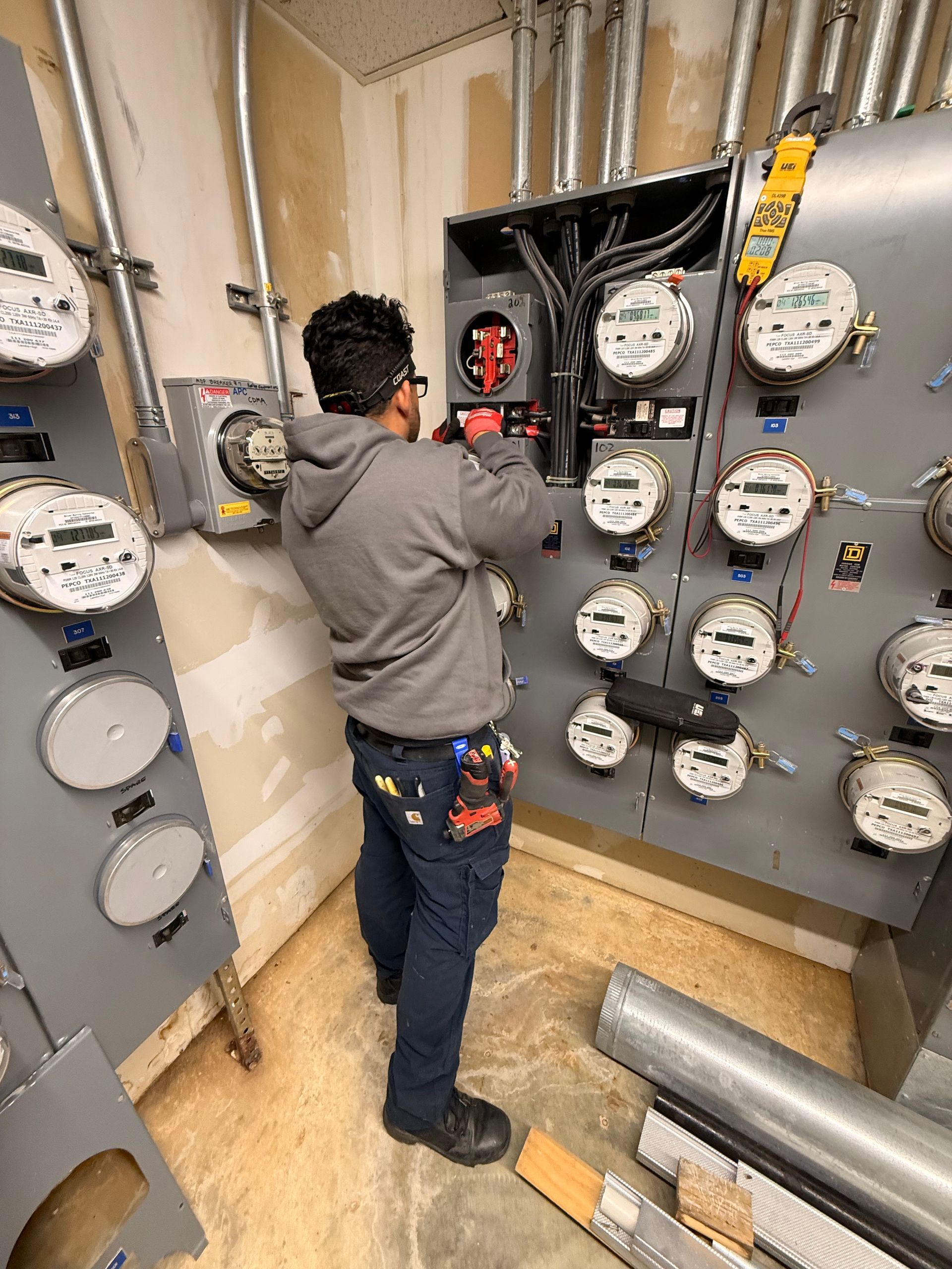 Electrician working on a gray electrical panel with multiple meters in an indoor setting.