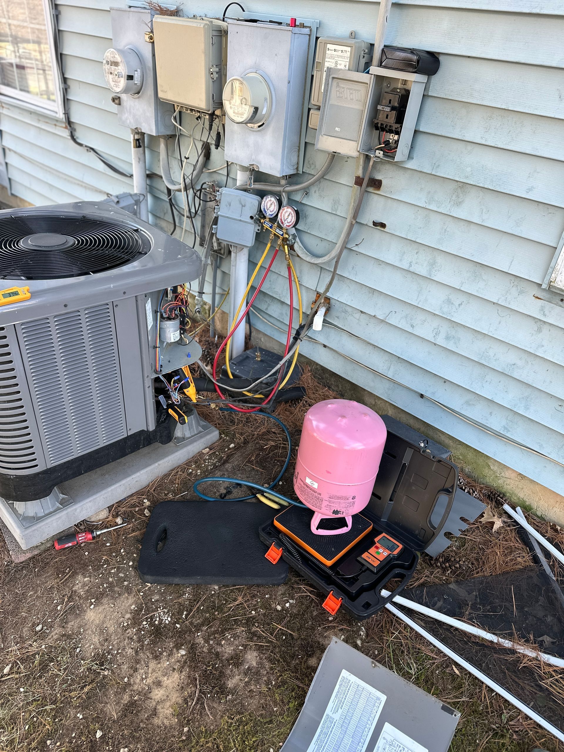 HVAC technician working on an AC unit outside a house; gauges, pink tank, tools, and electrical boxes are visible.