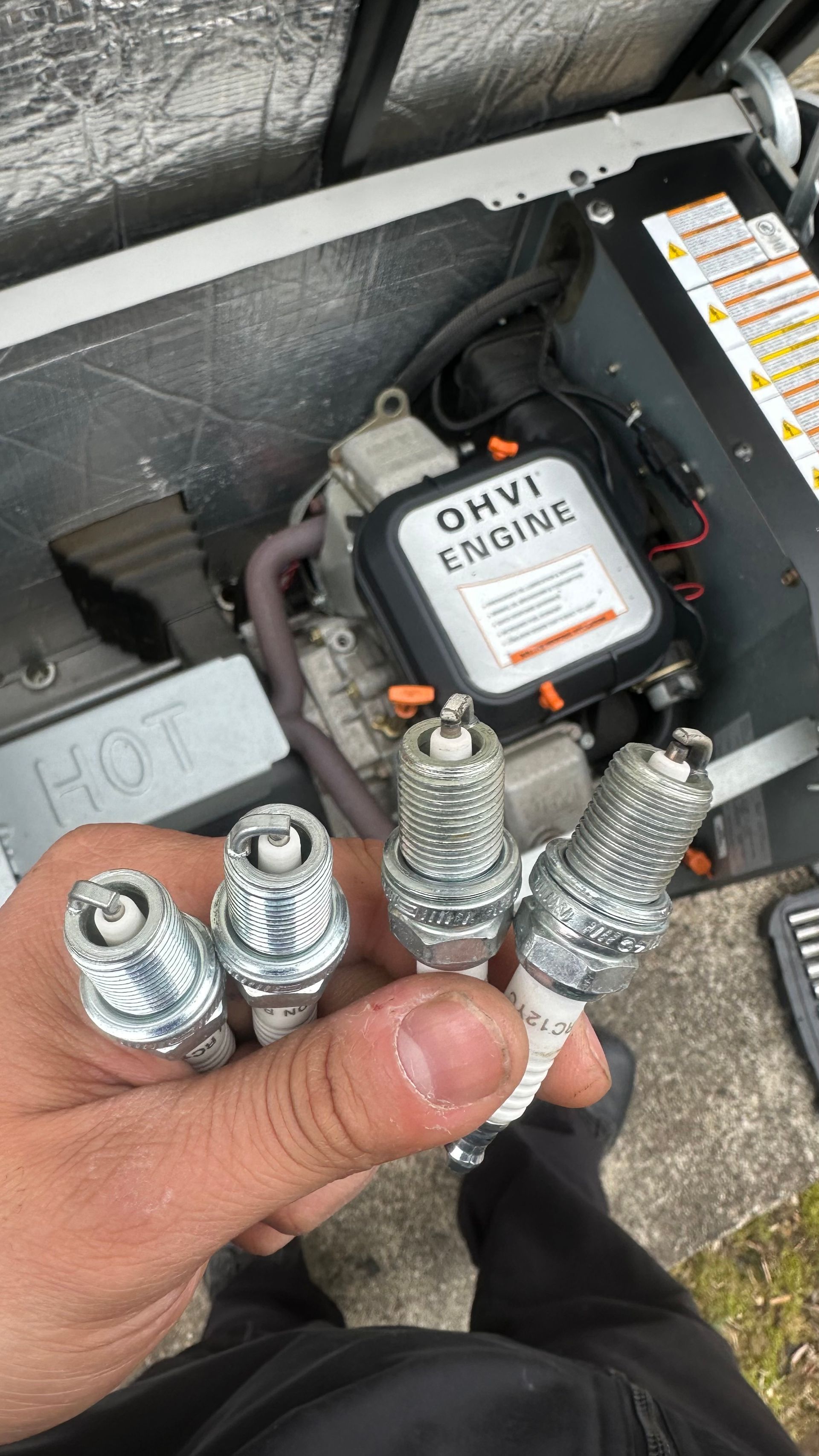Hand holding four used spark plugs in front of a generator with an OHV engine.