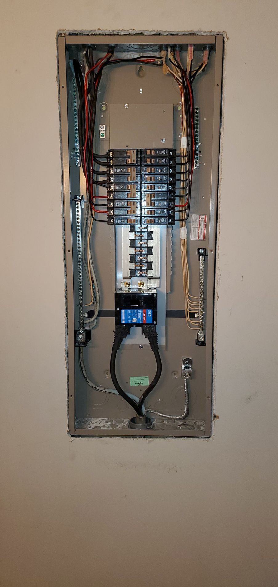 An open, gray electrical panel mounted on a beige wall with various wires and circuit breakers.