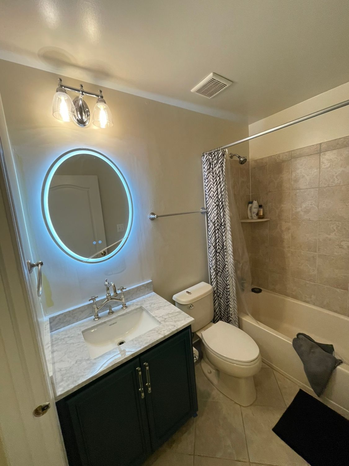 Bathroom with vanity, toilet, and bathtub/shower. Round mirror with blue light, teal vanity. Beige walls and tile.