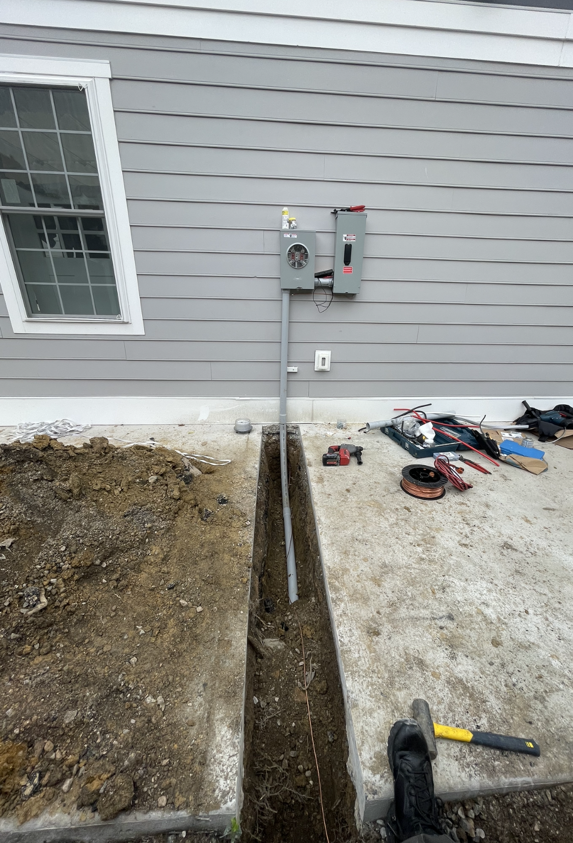 Trench dug next to a concrete walkway, with electrical conduit running up to a gray box on a house wall.