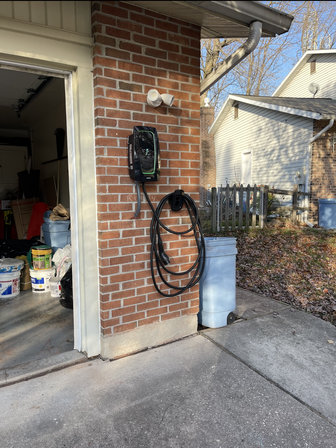 Electric vehicle charger mounted on a brick wall beside a garage door. A coiled charging cable hangs below.