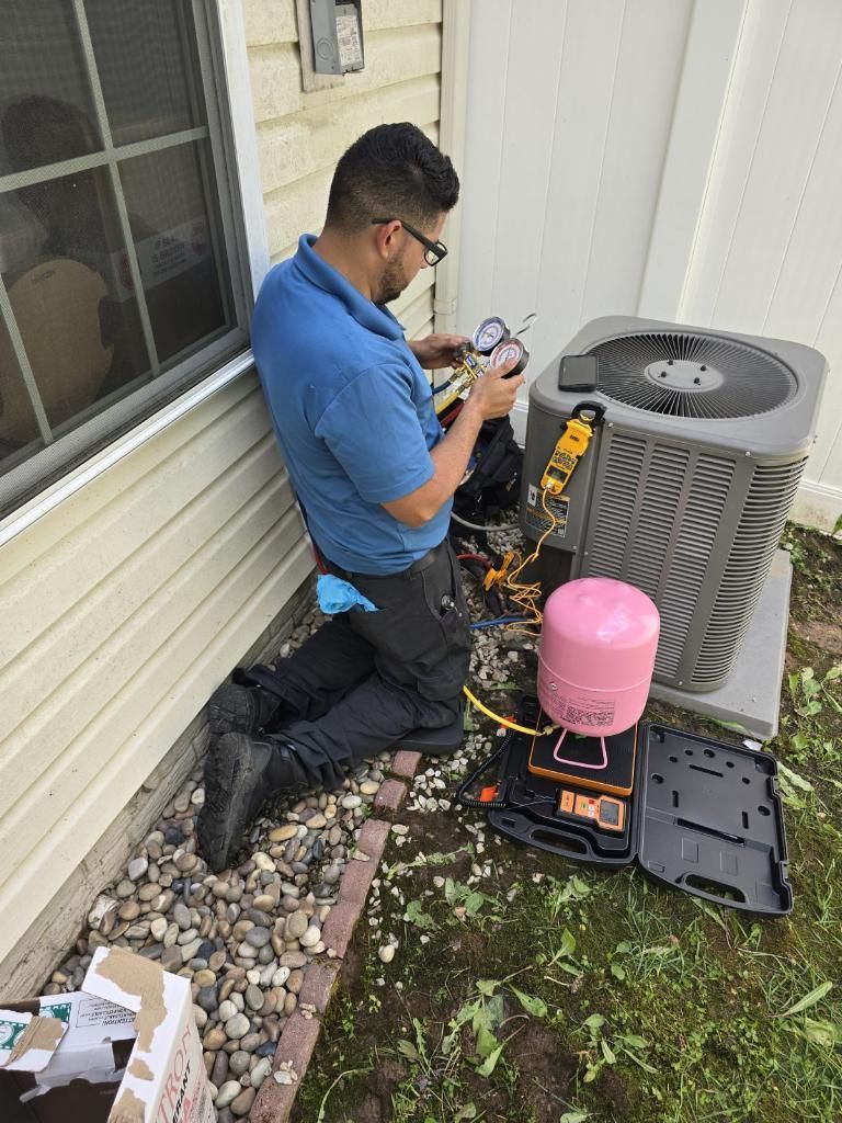 HVAC technician kneels near an AC unit, using gauges for maintenance. Outdoor setting, grass, tan siding.