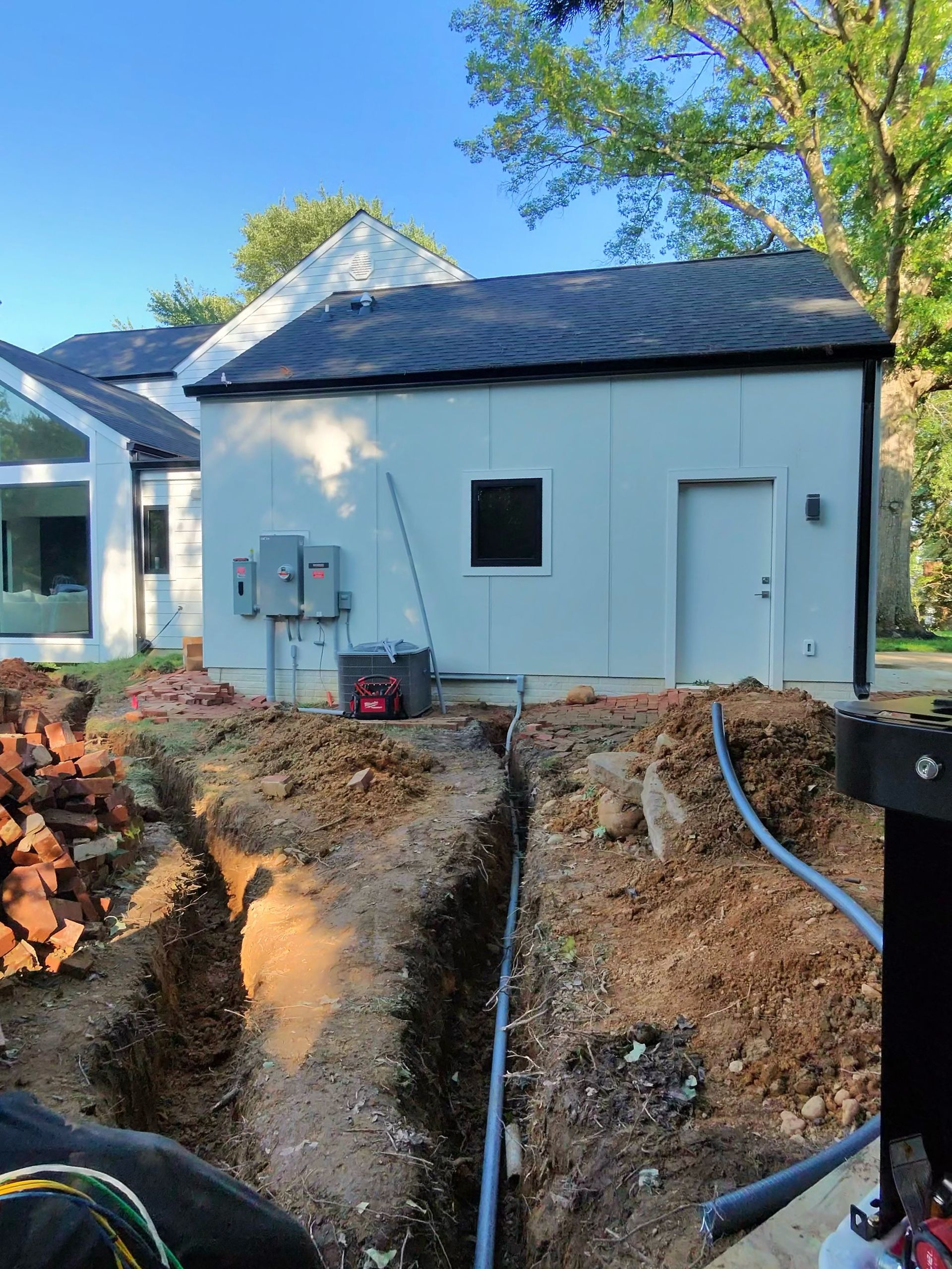 Construction site with a white building. A trench with a pipe runs towards the structure. Red bricks are piled nearby.