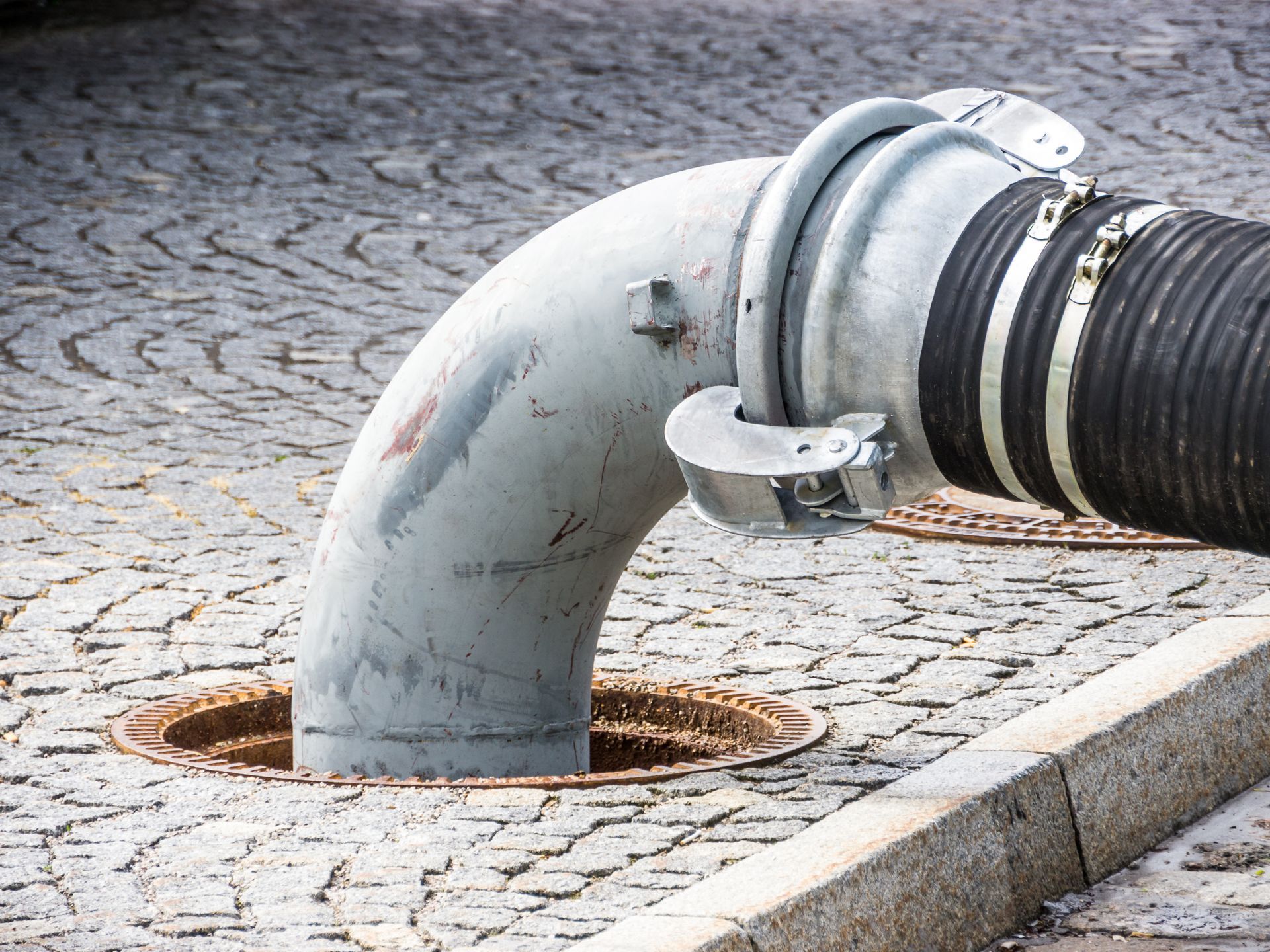 A large grey pipe bends into an open manhole on a cobblestone street.