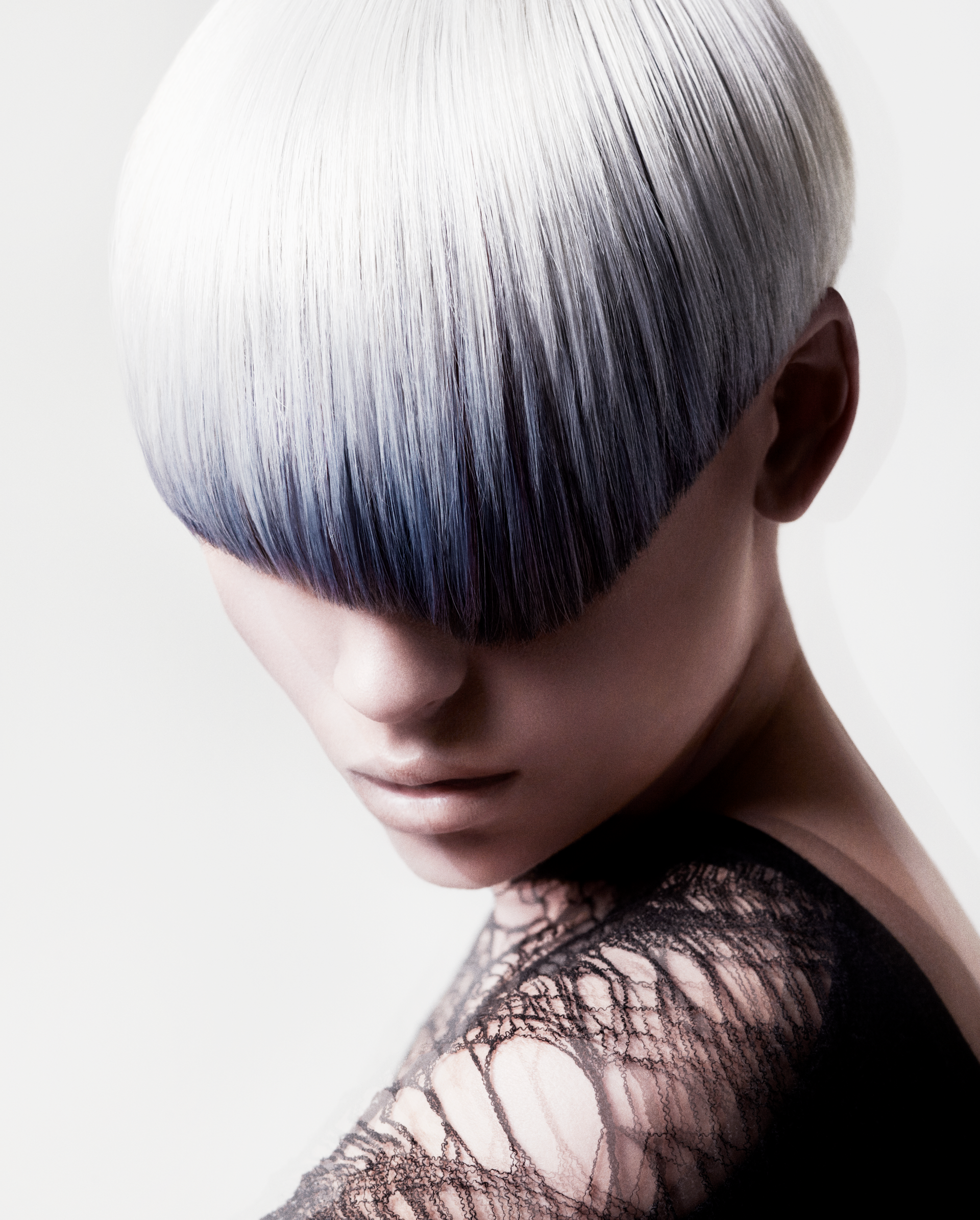 Woman with a bowl haircut, ombre of white to dark blue, looking down. She wears a black top and has a detailed tattoo on her shoulder.