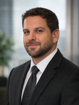 Man in a black suit and tie, standing in an office with a slight smile.
