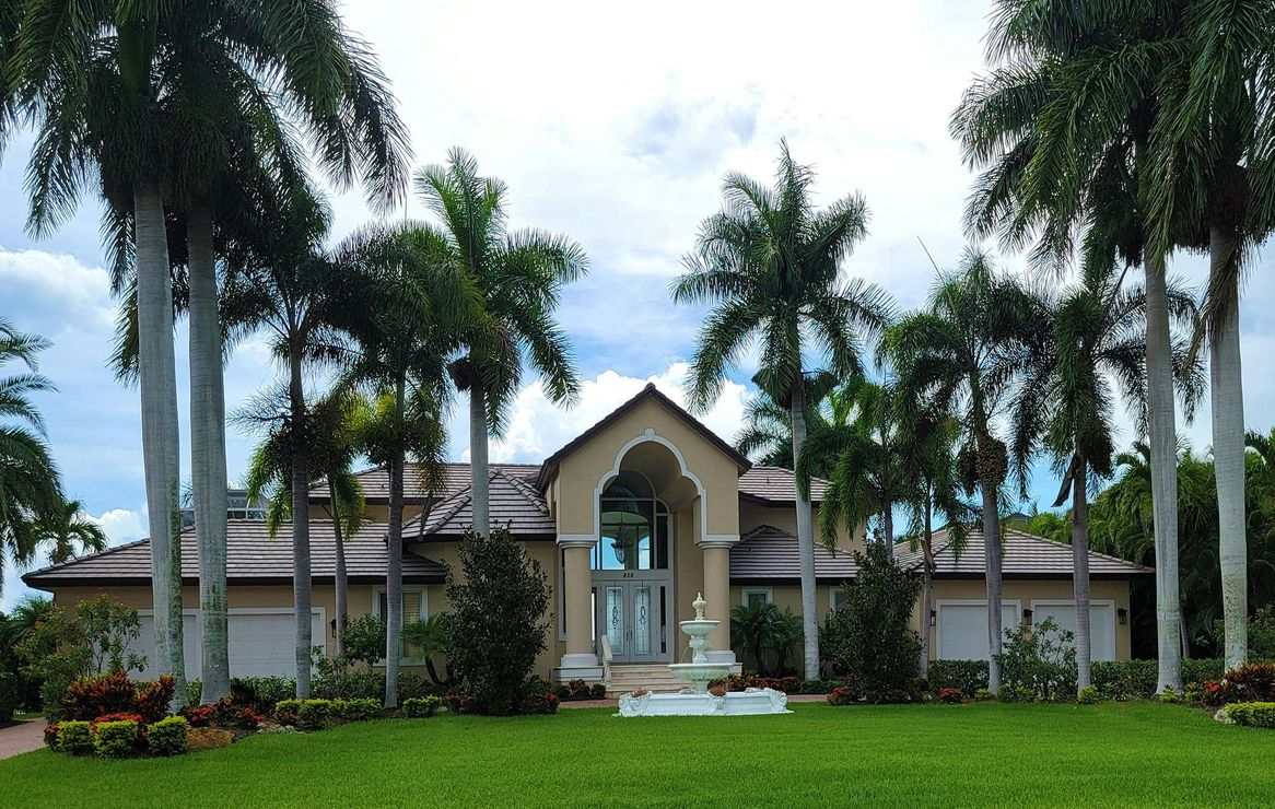 Tropical house with a central fountain, palm trees, and a lush green lawn under a blue sky