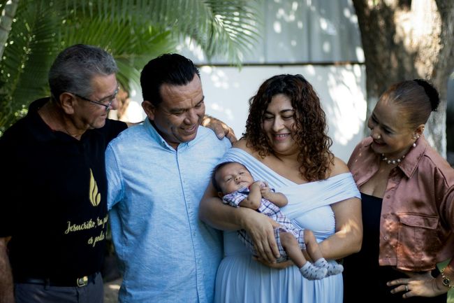 Five adults standing close together, smiling and holding a sleeping baby outdoors under palm leaves.