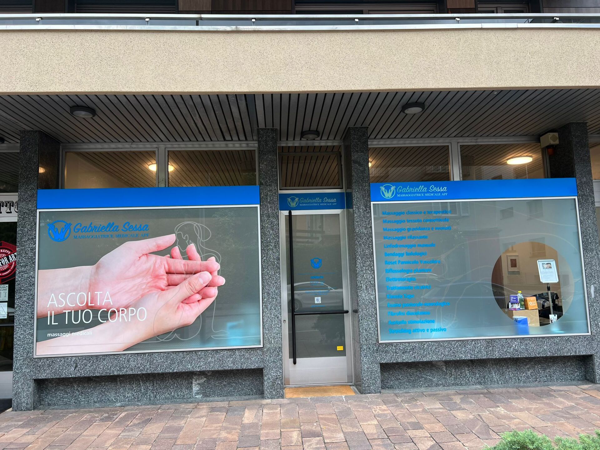Esterno della clinica audiologica; segnaletica blu e bianca; mani che tengono un apparecchio acustico.