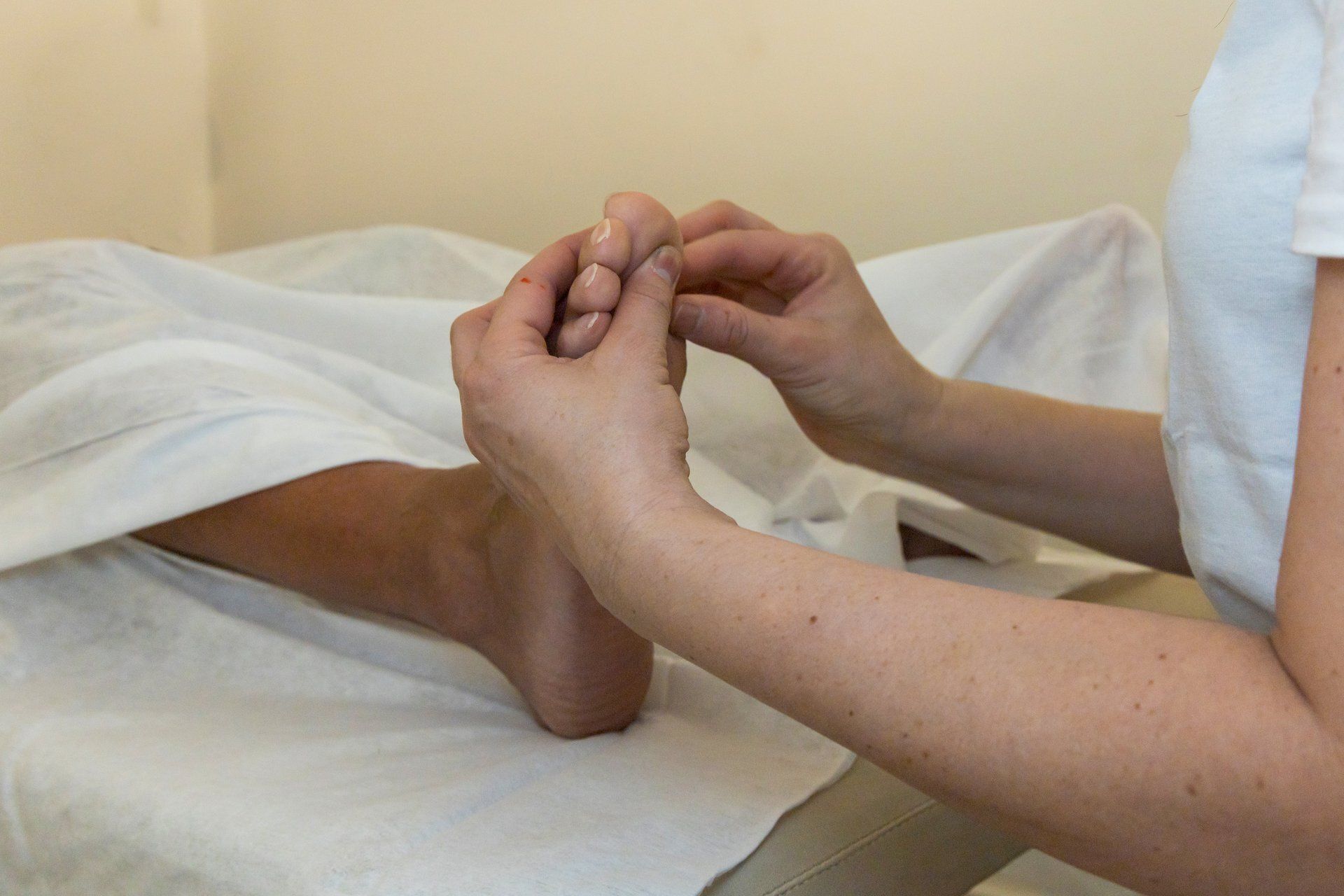 Mani che massaggiano un piede in un ambiente spa, concentrandosi sulle dita. Lenzuola e muro bianchi.
