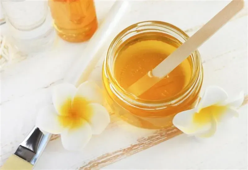 Open jar of amber honey with dipper on a bright white table, with plumeria flowers nearby