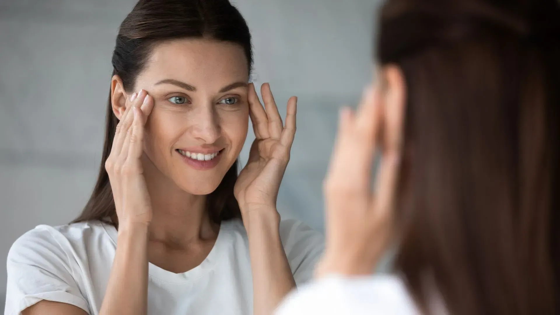 Woman smiling at a mirror while touching her face, with a bright reflection in a bathroom.