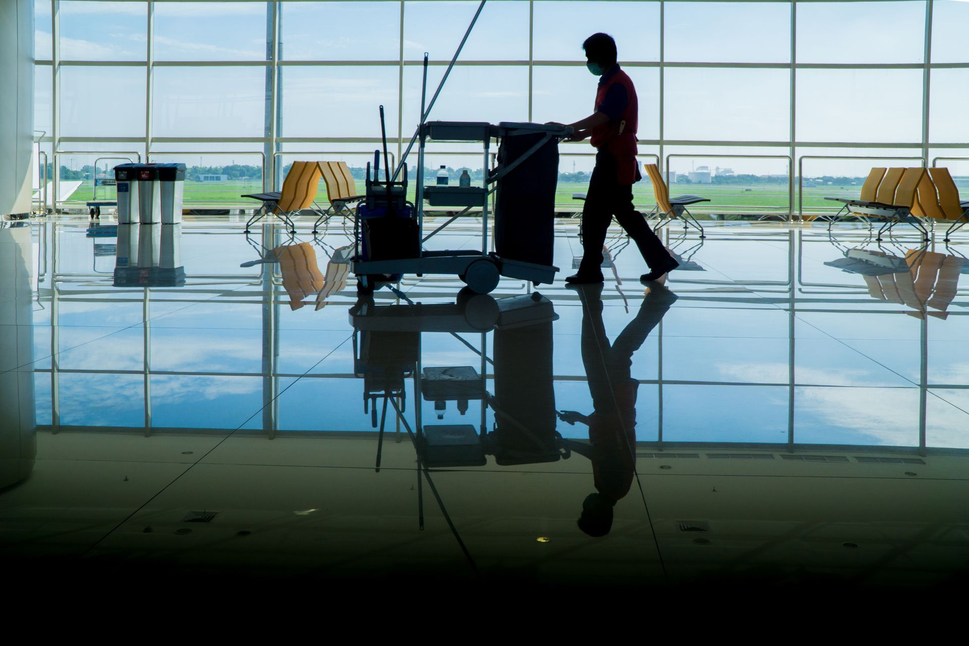 A silhouette of a worker pushing a cleaning cart across a polished floor in a bright, modern airport terminal.