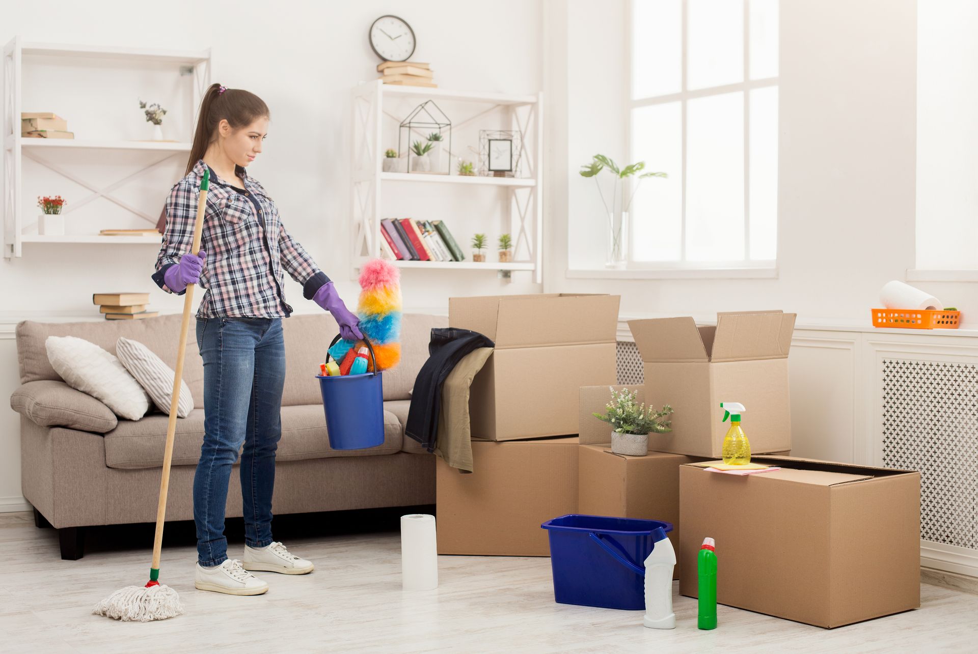 A person holds a mop and cleaning supplies in a living room filled with cardboard boxes, preparing to clean.