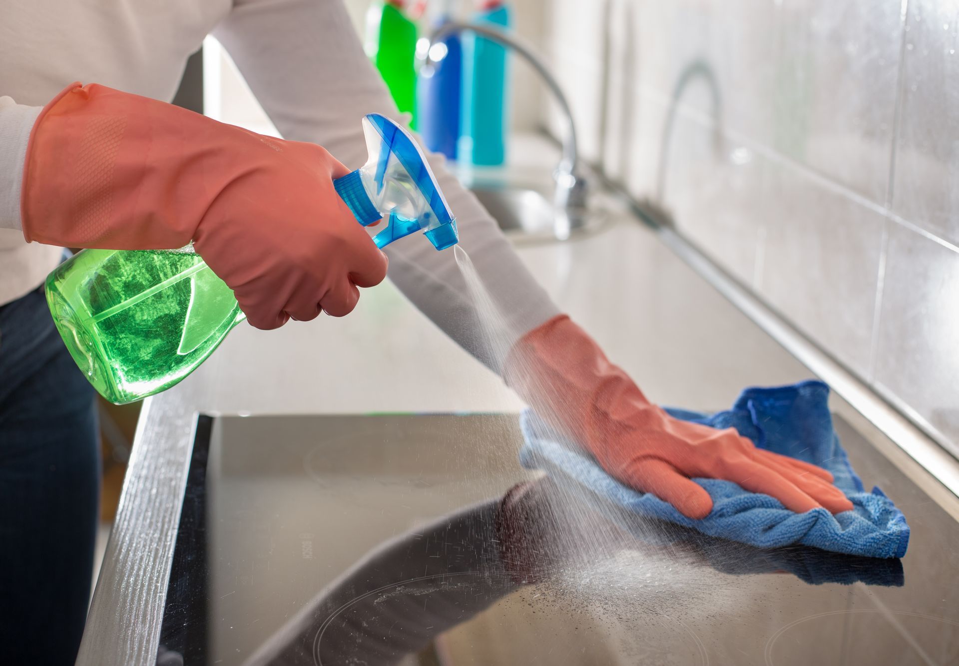 A person wearing orange rubber gloves uses a spray bottle and a blue cloth to clean a smooth, black kitchen surface.