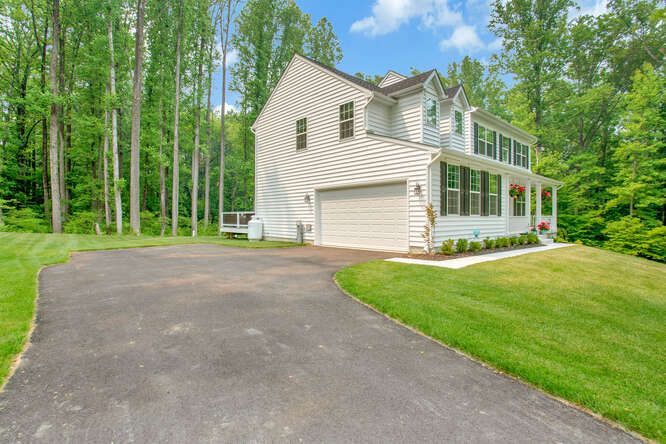 White house with black shutters, attached garage, and curved asphalt driveway on a grassy lot surrounded by trees.