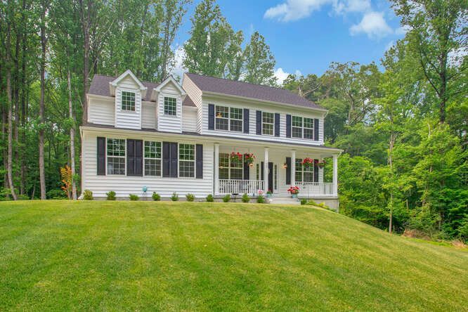 Two-story white house with black shutters and a front porch, on a green grassy hill, surrounded by trees.