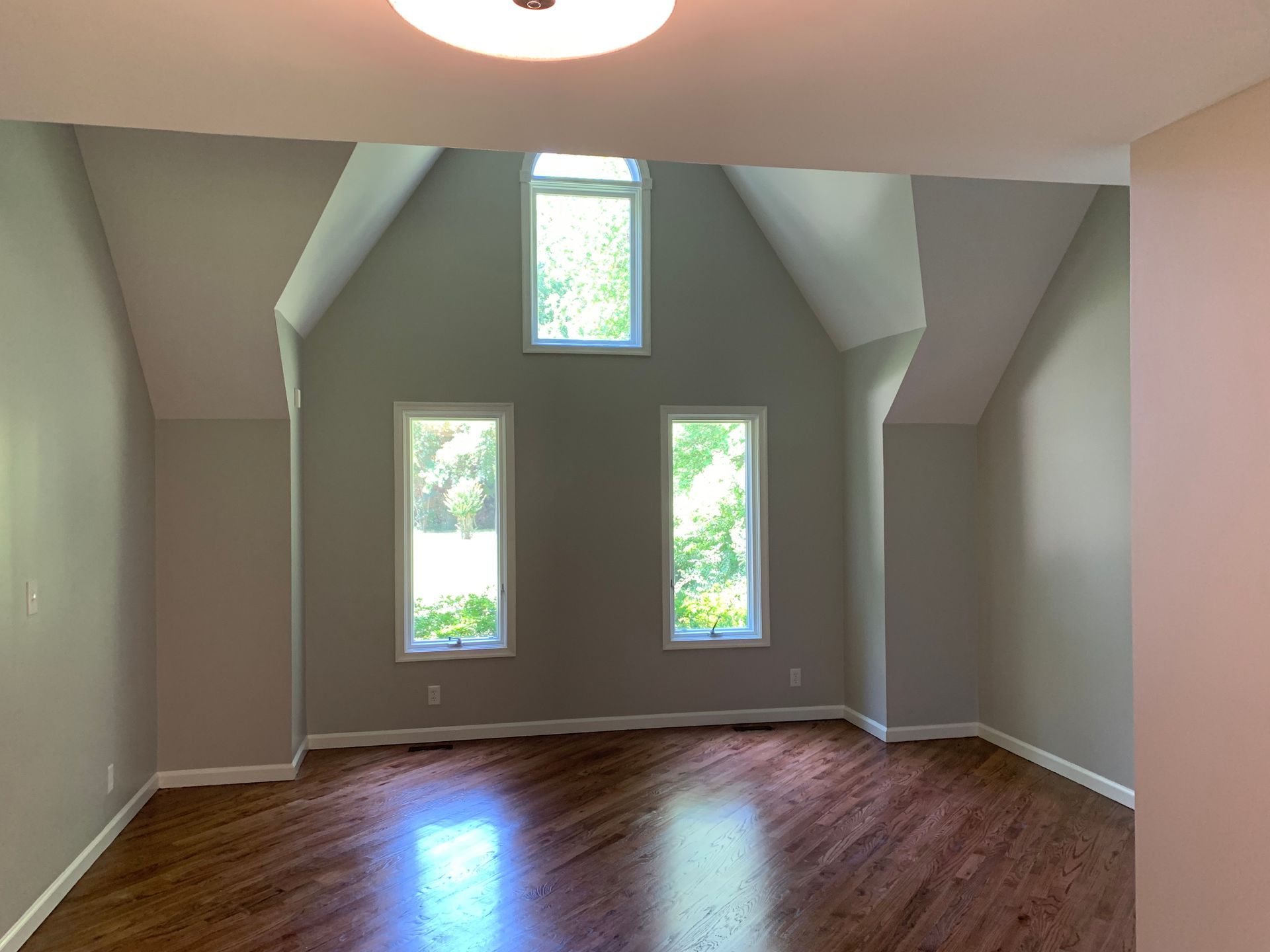 An empty room with wood flooring, light gray walls, and three windows, one arched, under a vaulted ceiling.