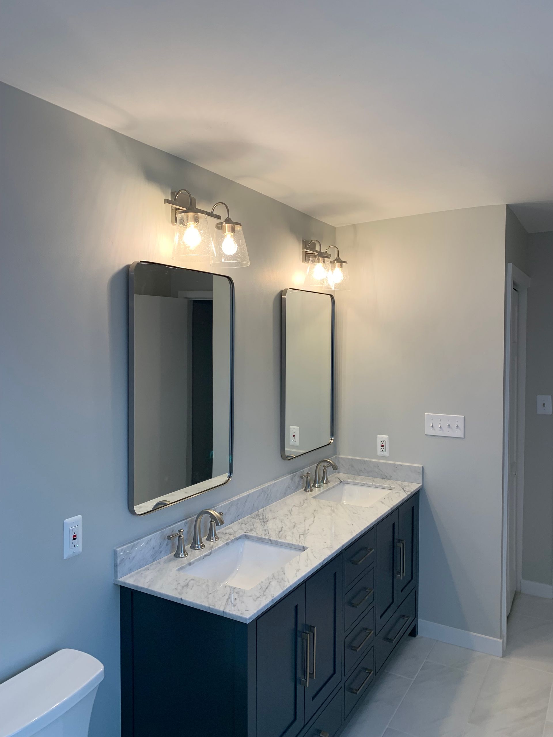 Bathroom with a dark blue vanity, two mirrors, and silver light fixtures. Light gray walls and white countertop.
