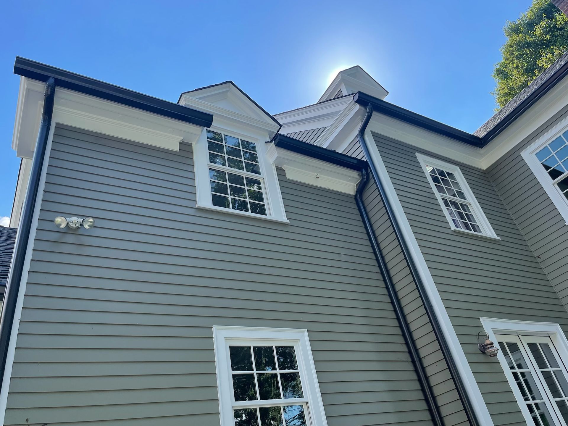 A house with gray siding, white trim, and black gutters against a bright blue sky. Sunlight streams from behind the roof.