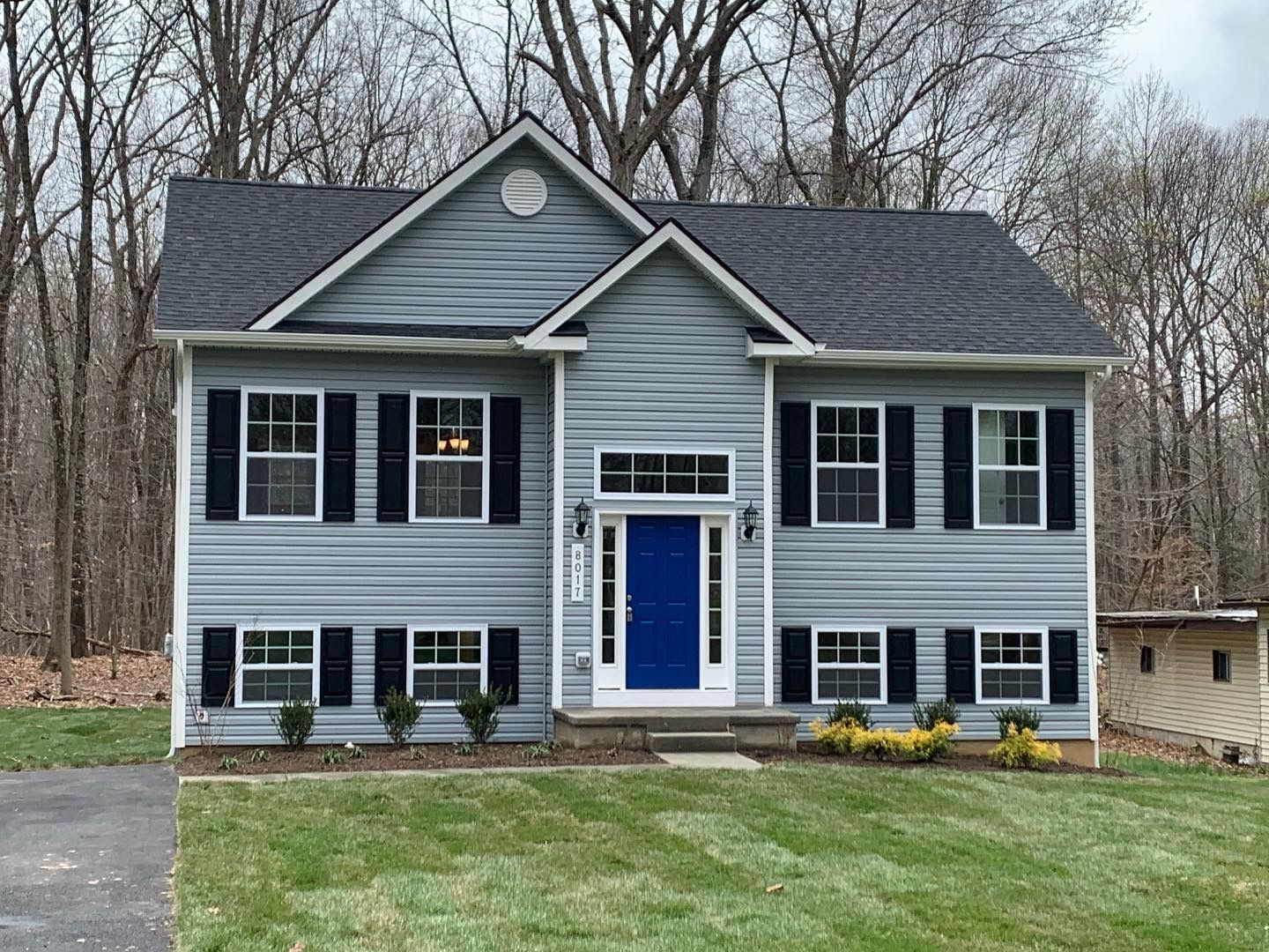 Blue-sided, two-story house with black shutters and a blue door in a yard surrounded by trees.