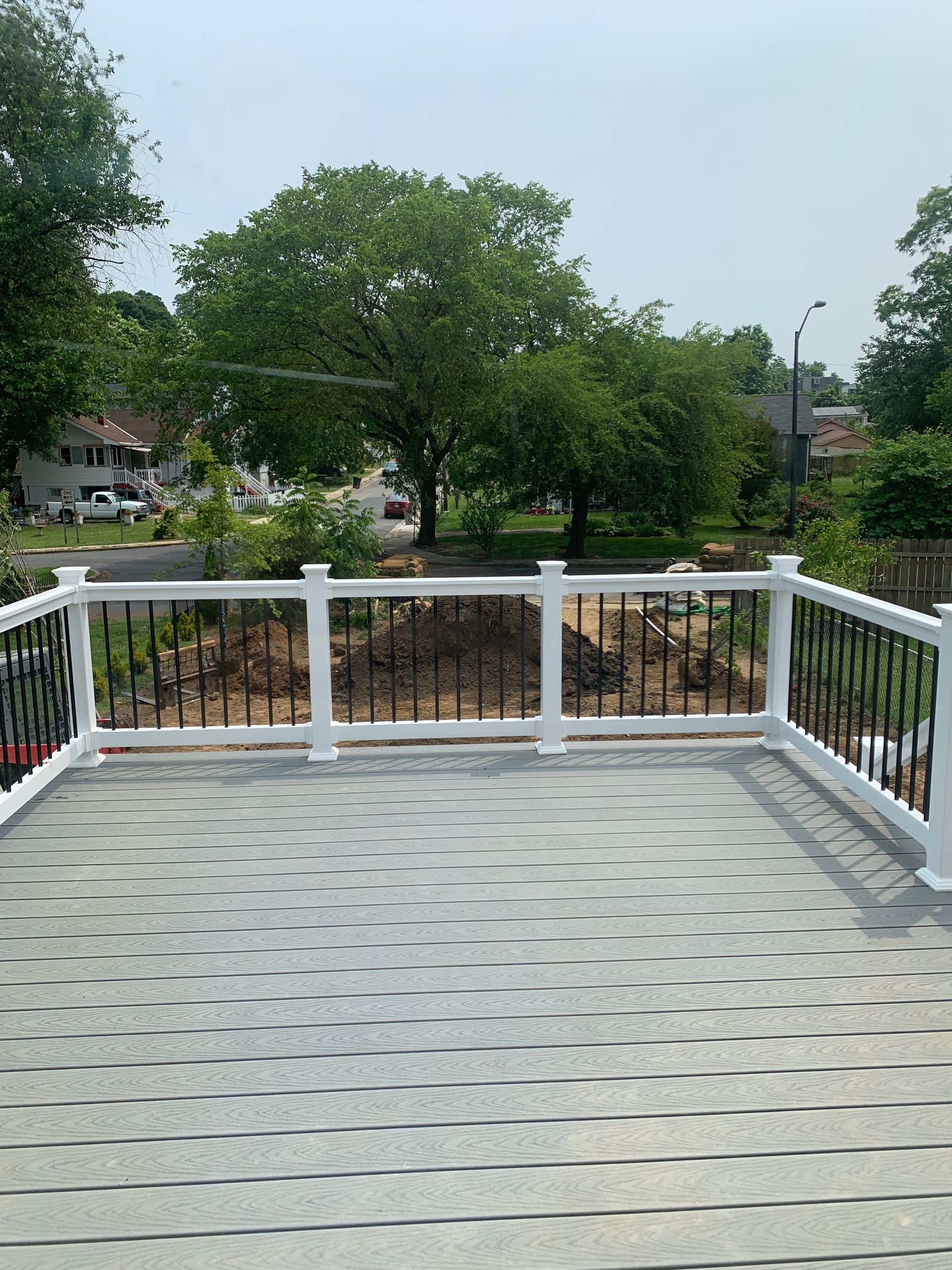 A gray deck with white posts and black railings overlooks a yard with trees and houses on a sunny day.