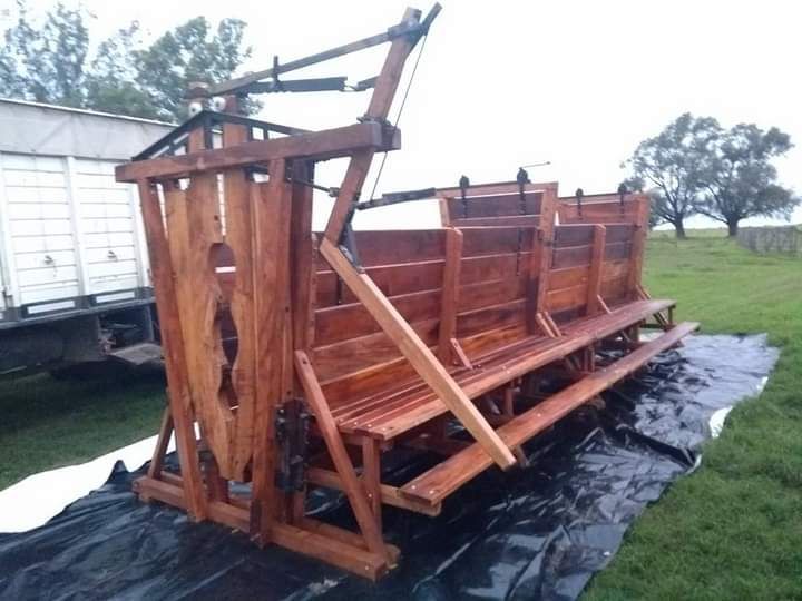 Conducto de madera para ganado con mecanismo de palanca, sobre una lona al aire libre, cerca de un camión y árboles.