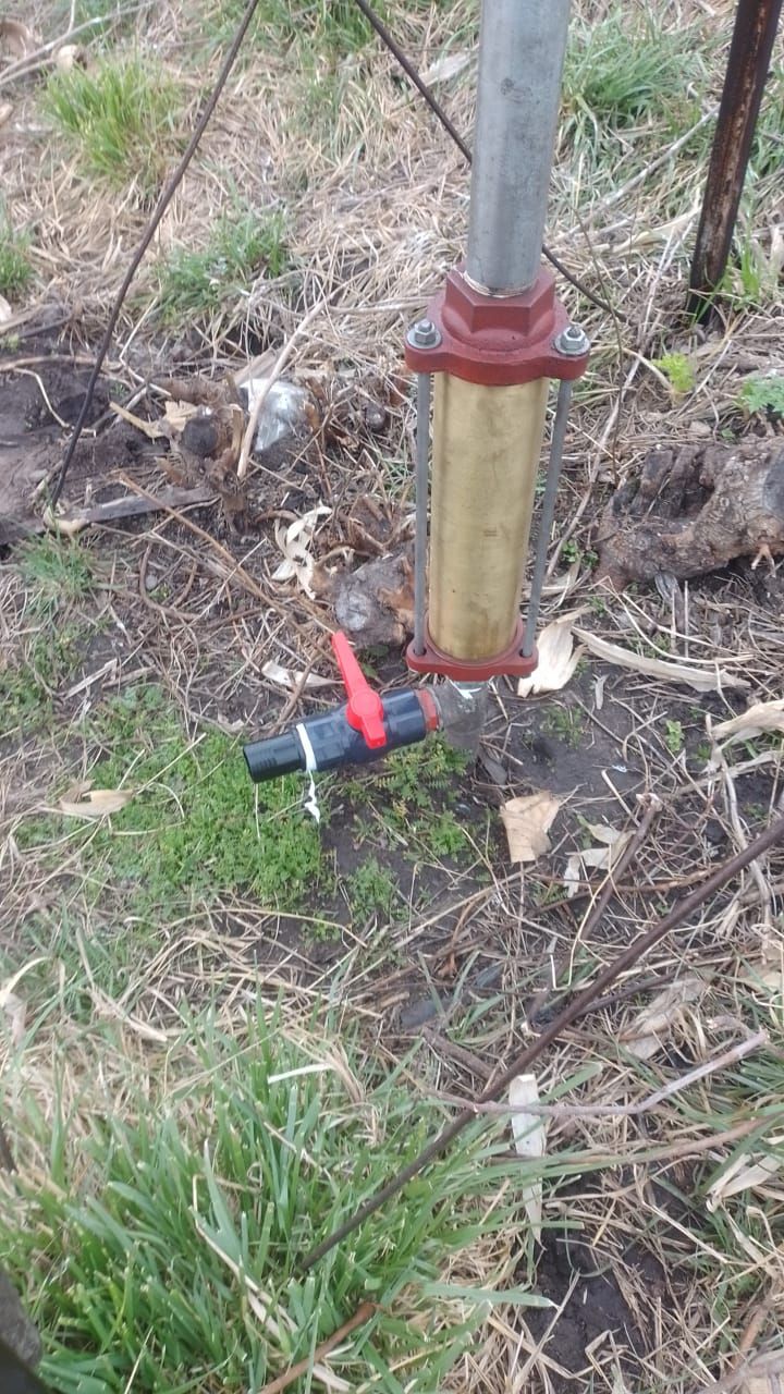 Una bomba de pozo con un cilindro de latón y una válvula roja cerca del suelo, en una zona cubierta de hierba.