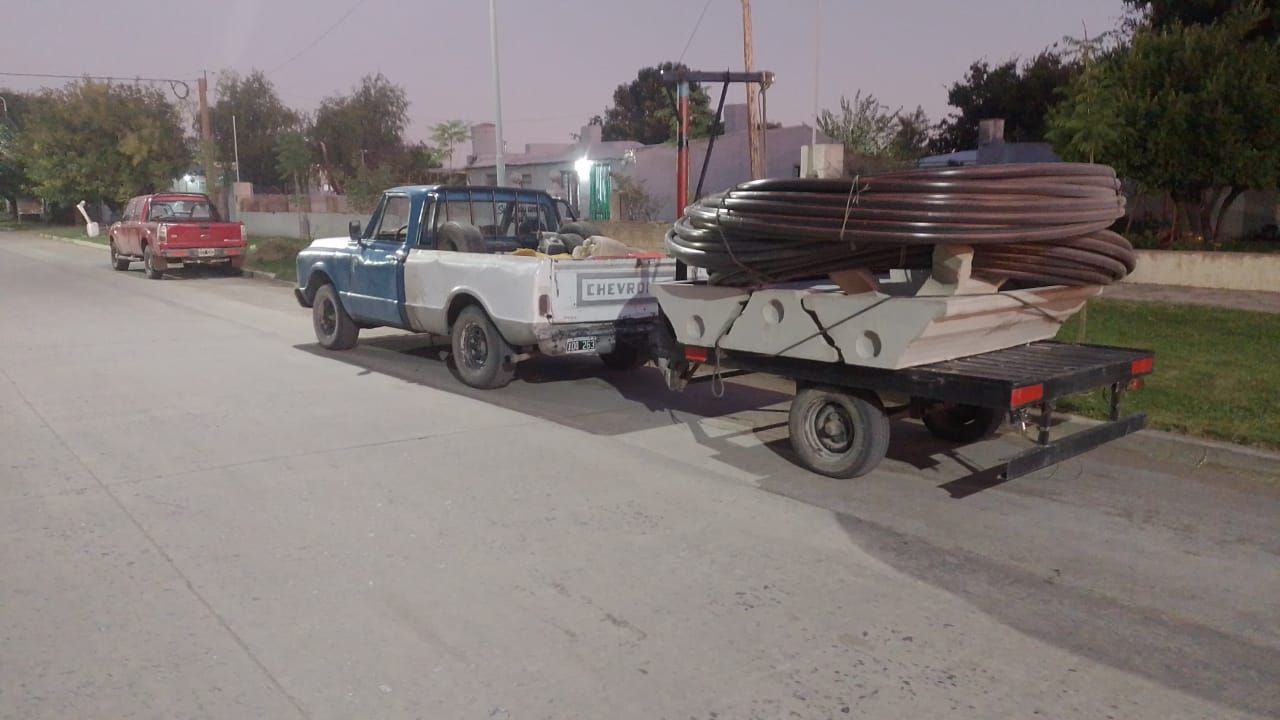 Una camioneta azul y blanca arrastraba un remolque cargado de tubos negros por la calle.