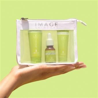 Hand holding a white, clear Image Skincare travel bag with green skincare products on a green background.