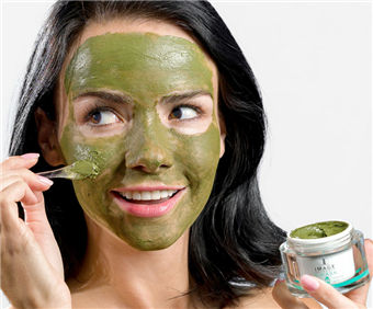 Woman with green face mask, smiling, applying more mask with a spatula. Holds jar of mask.