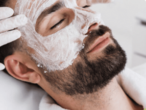 Man with a white facial mask being applied by gloved hands at a spa.