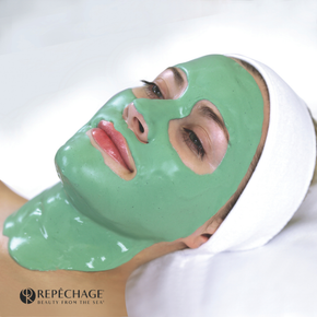 Woman with green face mask, smiling, applying more mask with a spatula. Holds jar of mask.