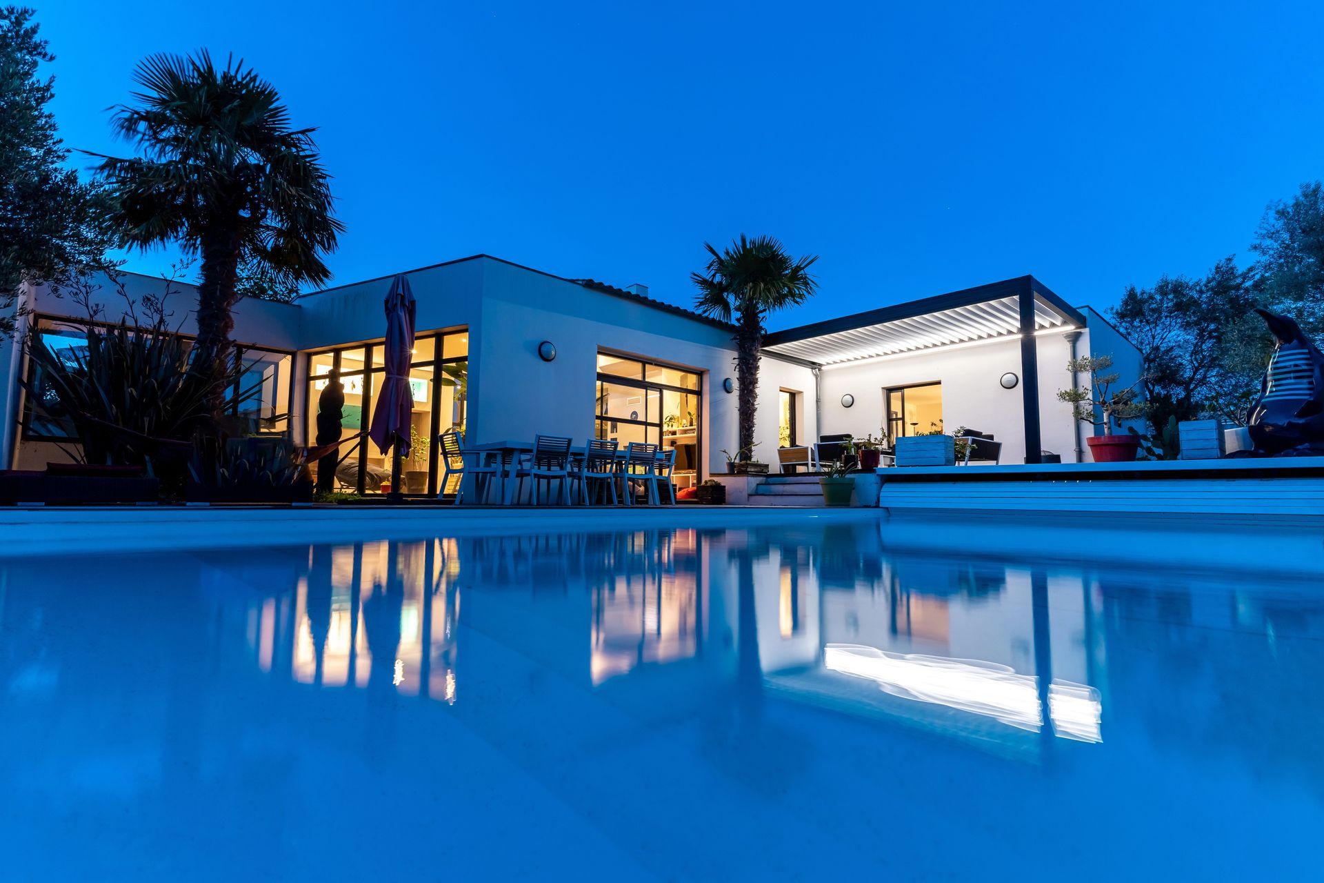 Una moderna casa bianca a un solo piano con finestre illuminate e un patio, che si riflette in una piscina al crepuscolo.