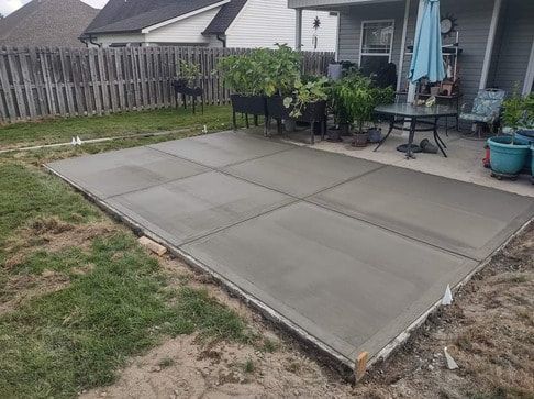 Concrete patio with square slabs beside a fenced backyard and patio furniture, next to a house.