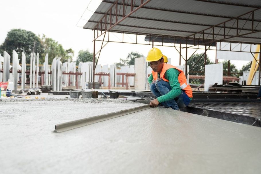 Construction worker smoothing wet concrete on a slab under a metal roof, wearing a yellow hard hat and orange vest