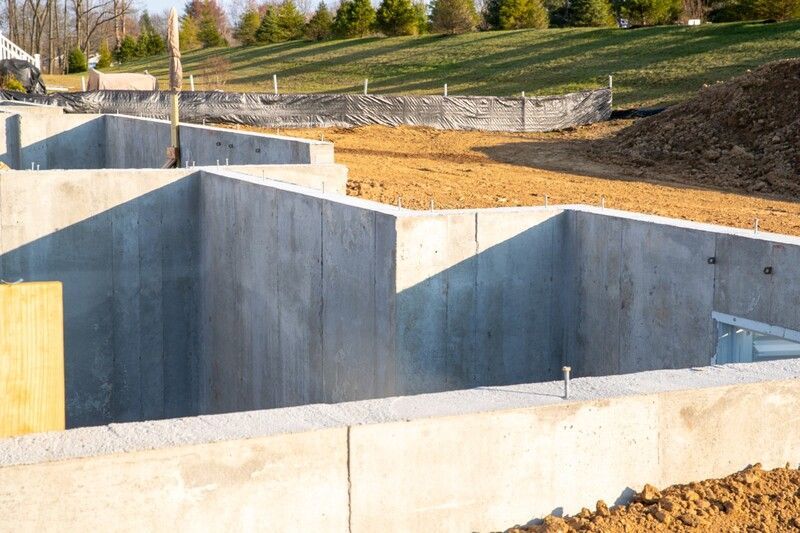 Concrete foundation walls under construction on a dirt lot, with grassy slope and trees in the background