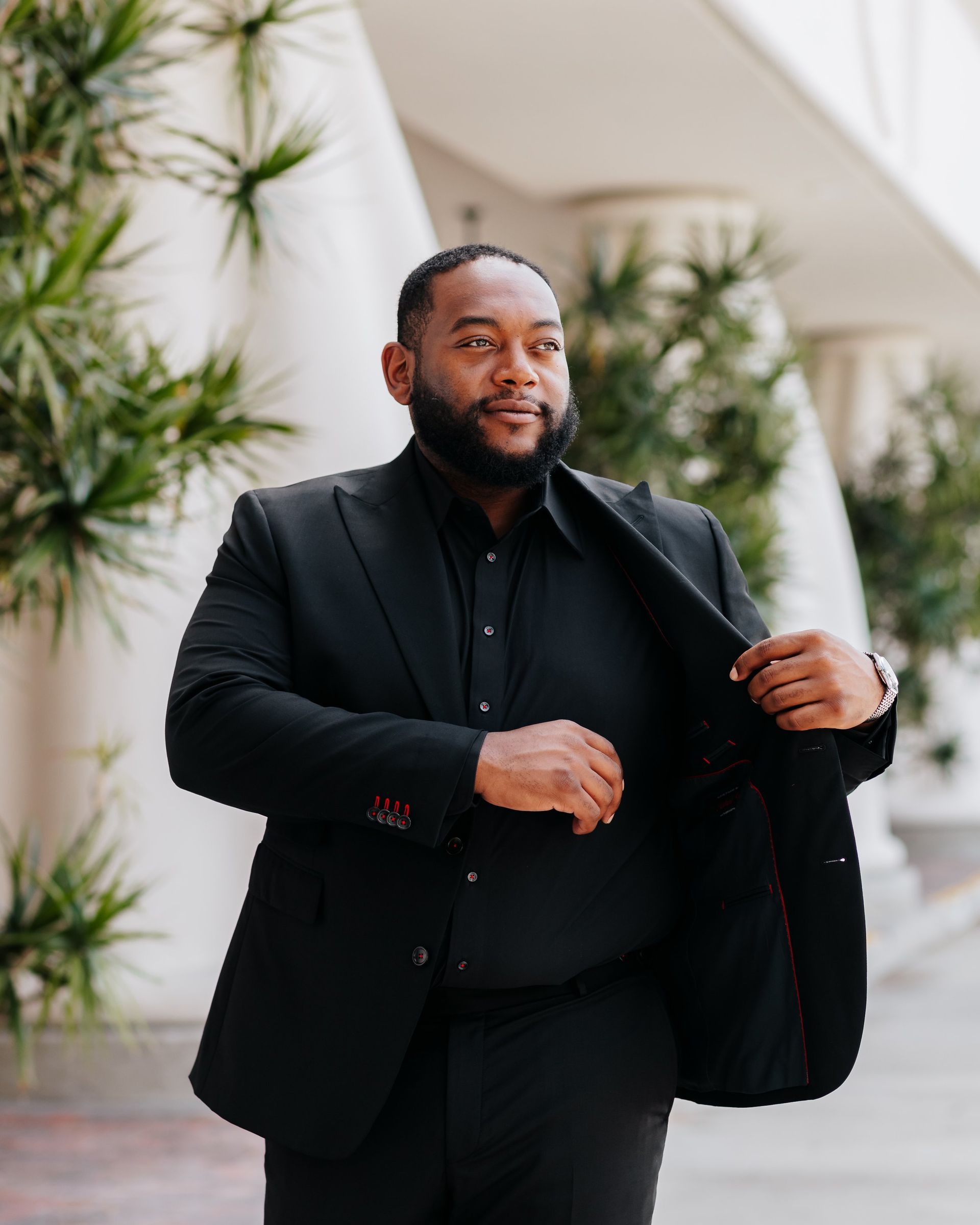 A man in a black suit and black shirt is standing in front of a building.