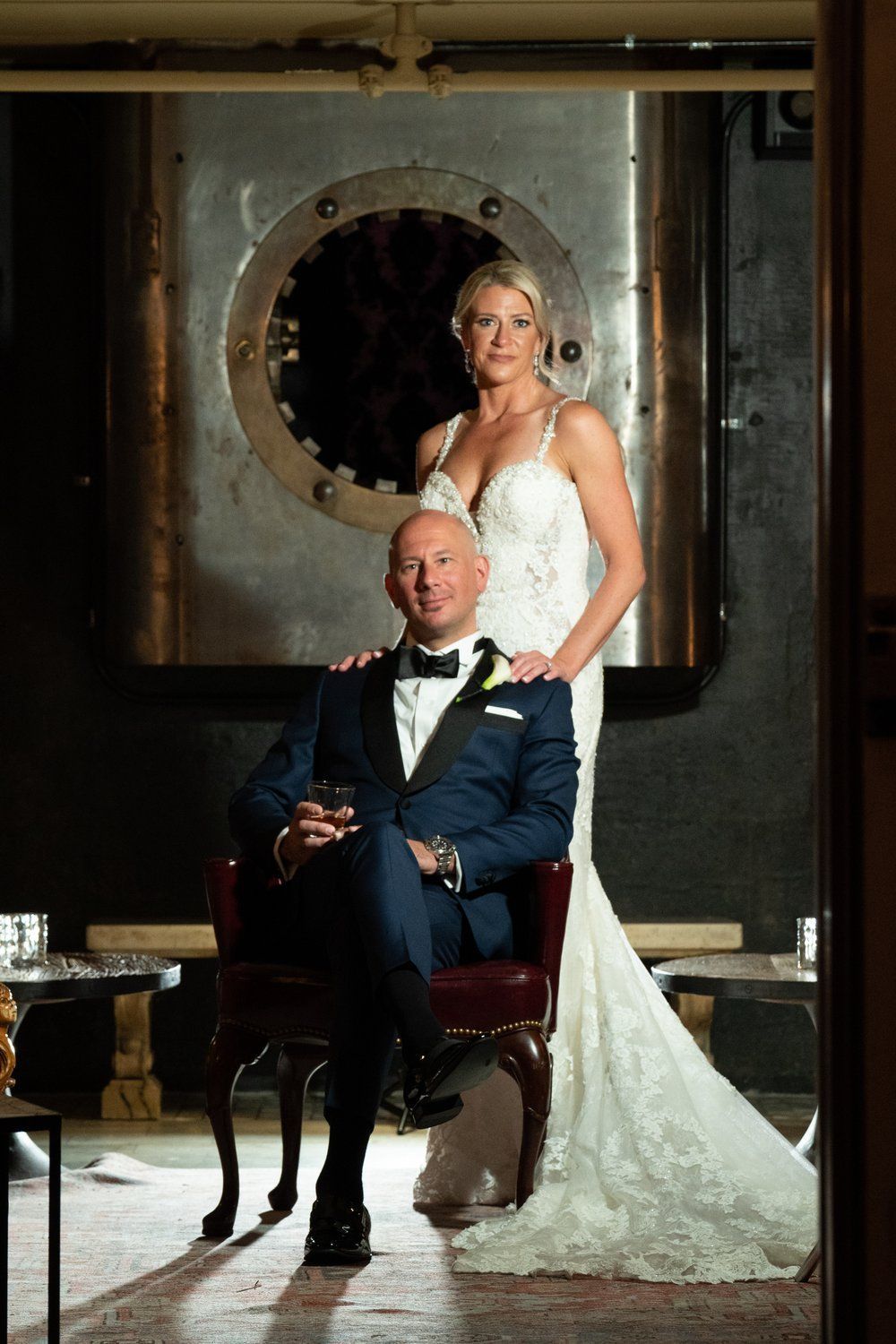 The bride and groom are posing for a picture while the groom is sitting in a chair.