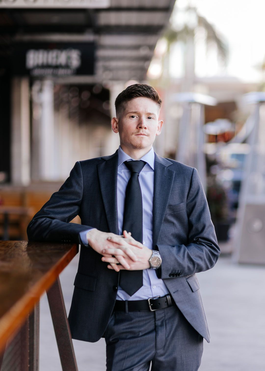 Man in a suit leans on a bar, looking directly at the camera. Outdoor setting, neutral colors.