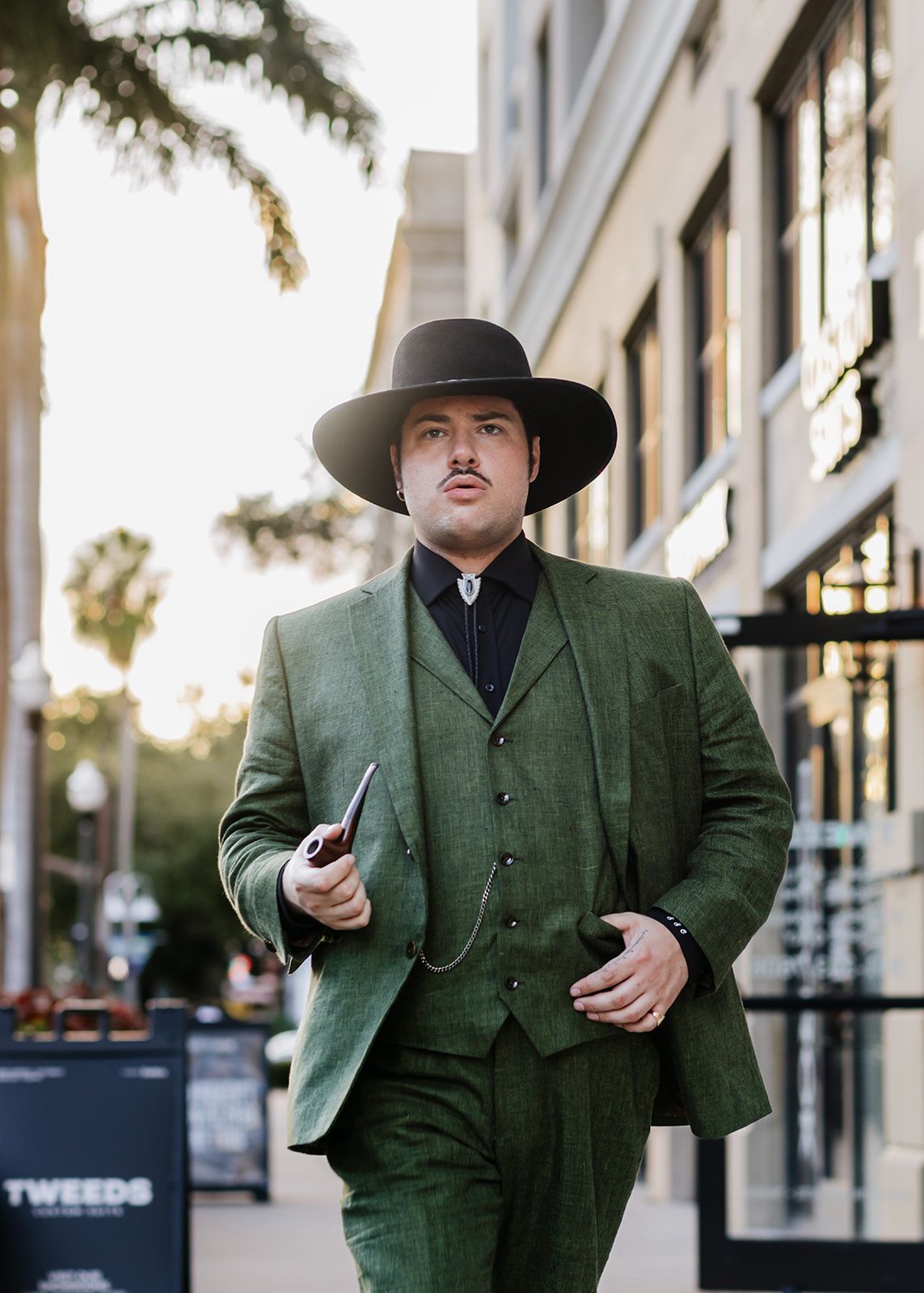 Man in a suit leans on a bar, looking directly at the camera. Outdoor setting, neutral colors.