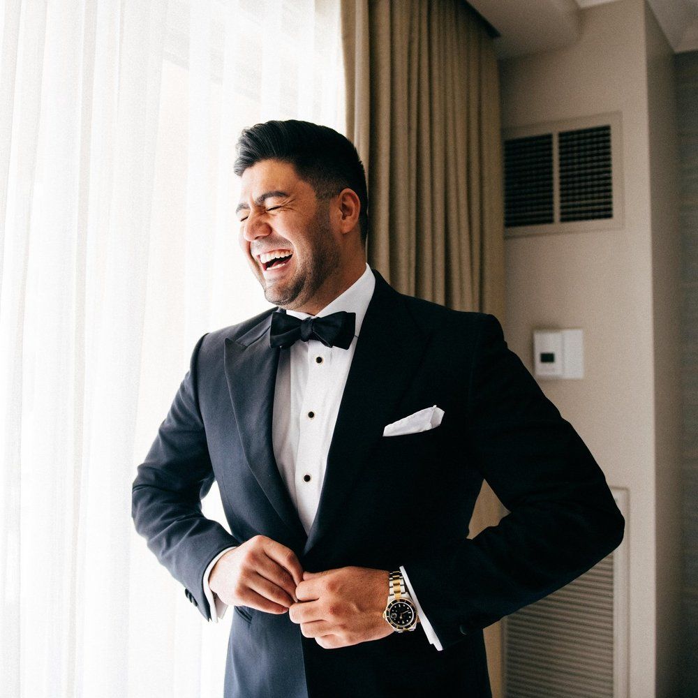 A man in a tuxedo is laughing in front of a window.